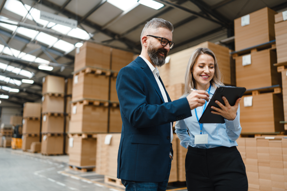 Man and woman in business attire reviewing a tablet together in a warehouse filled with stacked cardboard boxes.