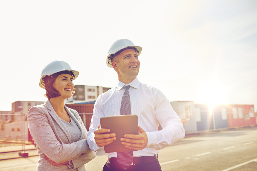 Two safety professionals wearing white hard hats discussing work outdoors with one holding a tablet, with buildings in the background and sunlight.