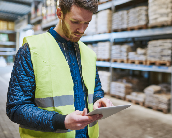 Man in a yellow safety vest looking at a tablet inside a warehouse with shelves of stacked bags.