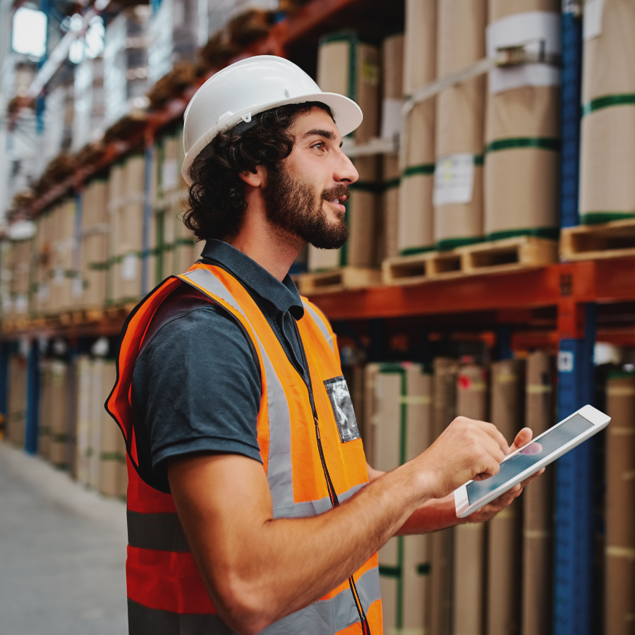 Warehouse worker wearing a white hard hat and orange safety vest using a tablet in front of stacked cardboard rolls on shelves.