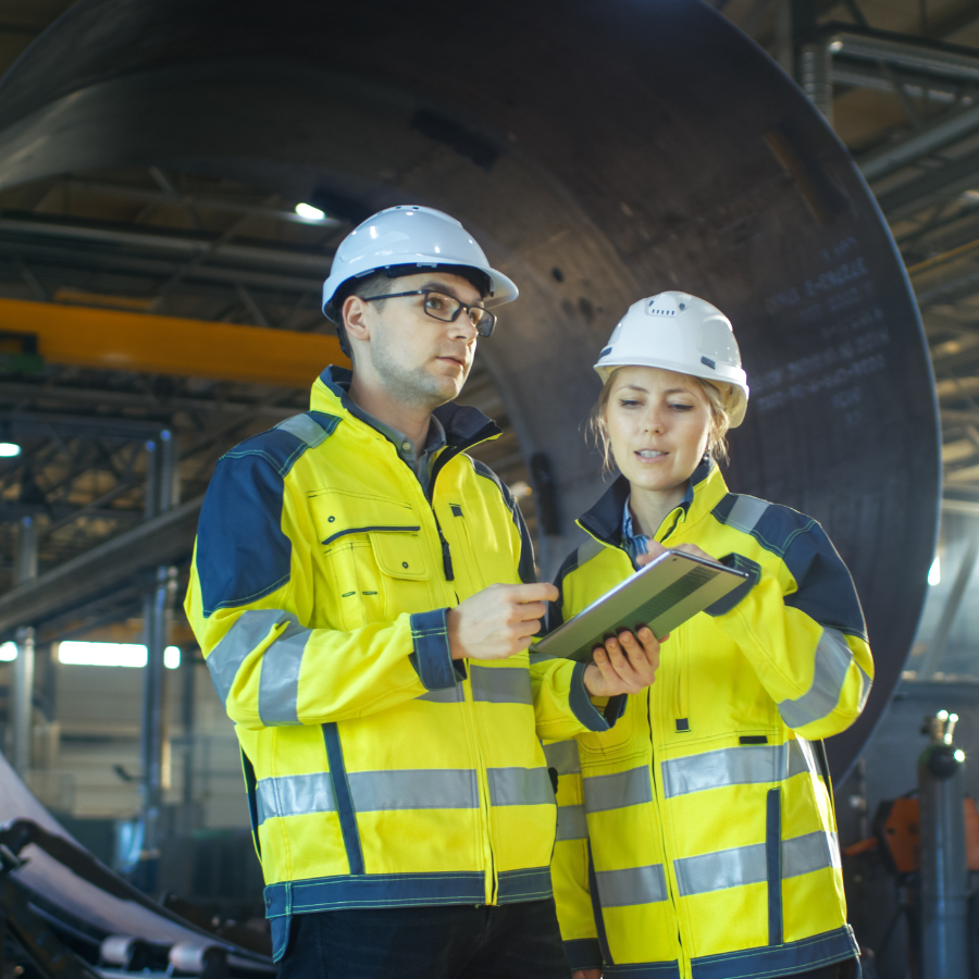 Two industrial workers in yellow reflective jackets and white hard hats discussing information on a clipboard inside a factory.