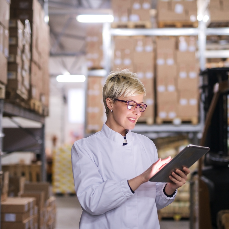 Smiling woman in a white lab coat using a tablet in a warehouse with stacked boxes.