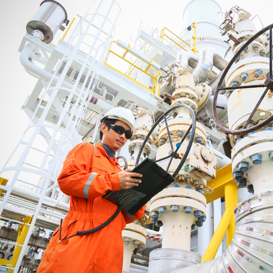 Worker in orange safety uniform and helmet using a tablet near industrial pipes and valves.