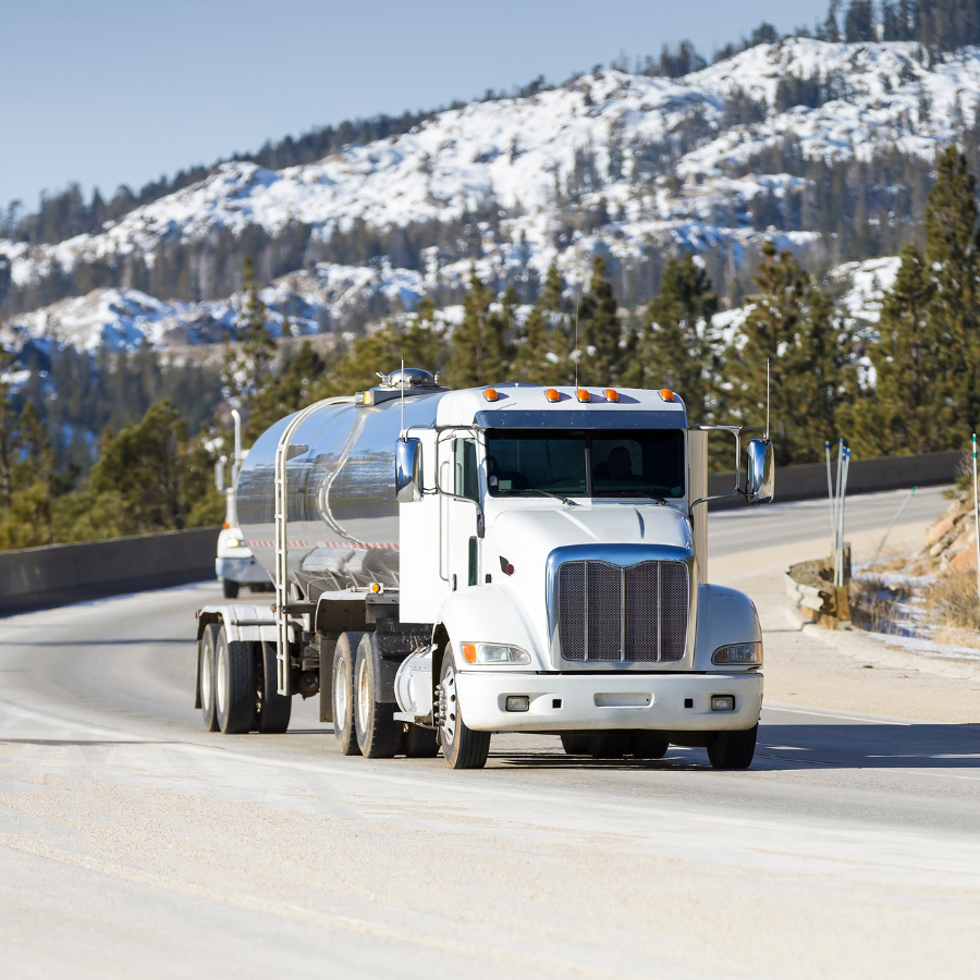 White tanker truck driving on mountain road with snow-covered trees and hills in the background.