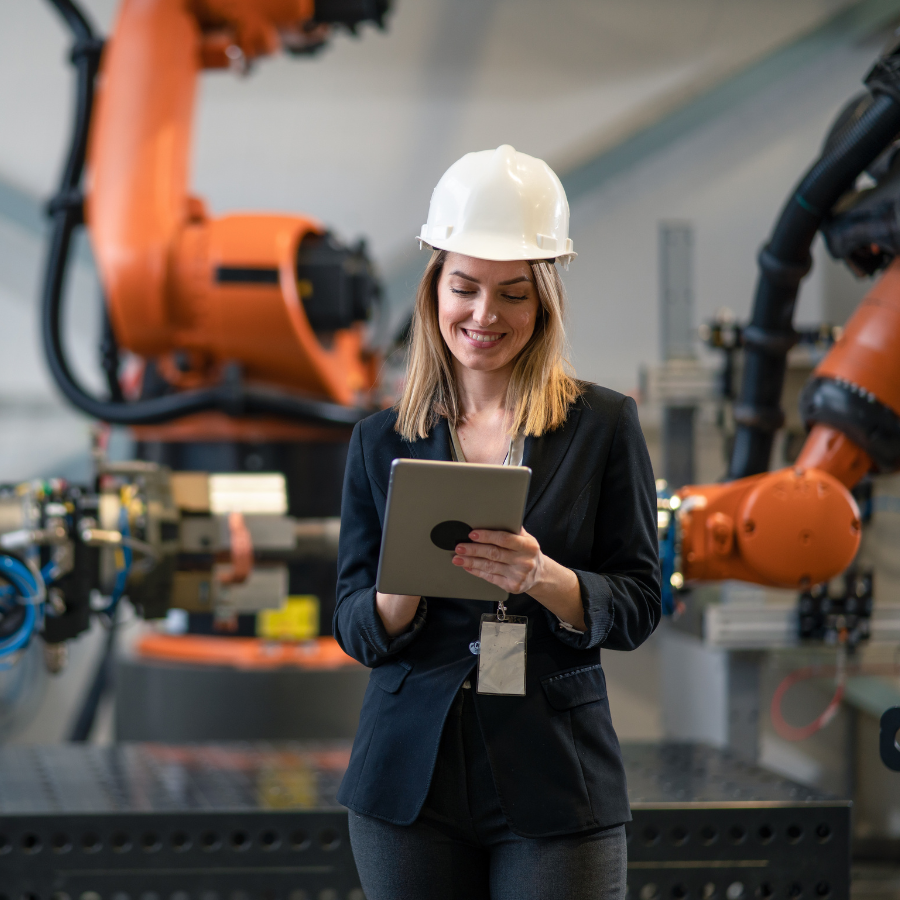 Smiling female engineer wearing a white hard hat and black blazer using a tablet with industrial robotic arms in the background.