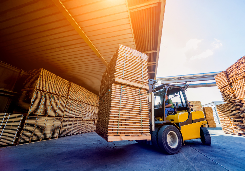 Yellow forklift lifting a large stack of wooden planks in a lumber yard with more stacked wood under a roof.
