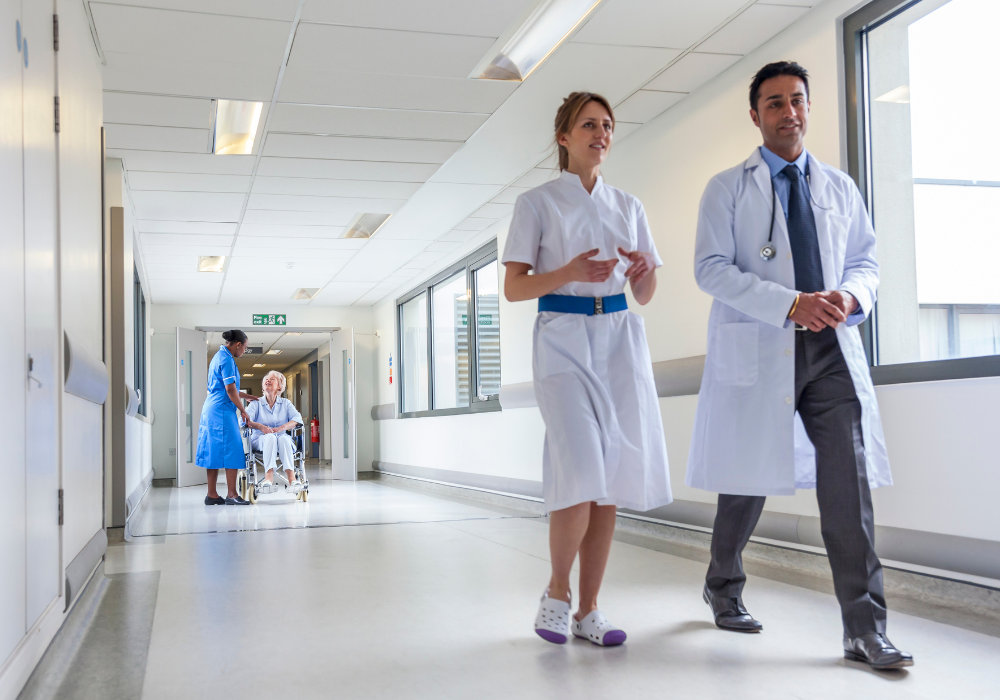 Two healthcare professionals walking down a hospital corridor as a nurse assists an elderly patient in a wheelchair in the background.