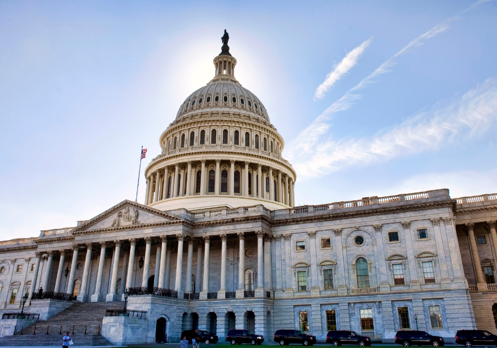 United States Capitol building with its dome and a flagpole, under a clear blue sky with sunlight behind the dome.