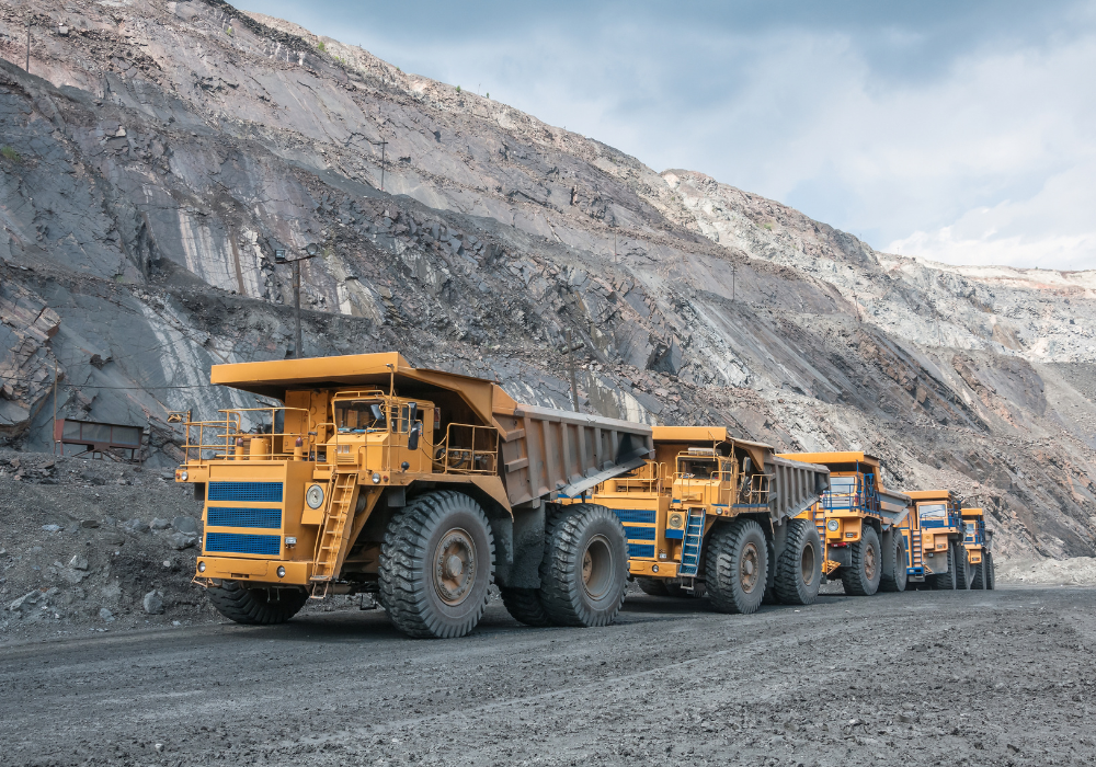 Four large yellow mining dump trucks lined up on a dirt road in an open-pit mine with rocky cliffs in the background.