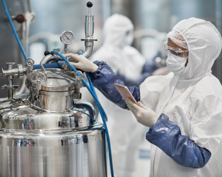 Scientist in full protective suit, gloves, and mask operating a stainless steel chemical reactor and holding a tablet.