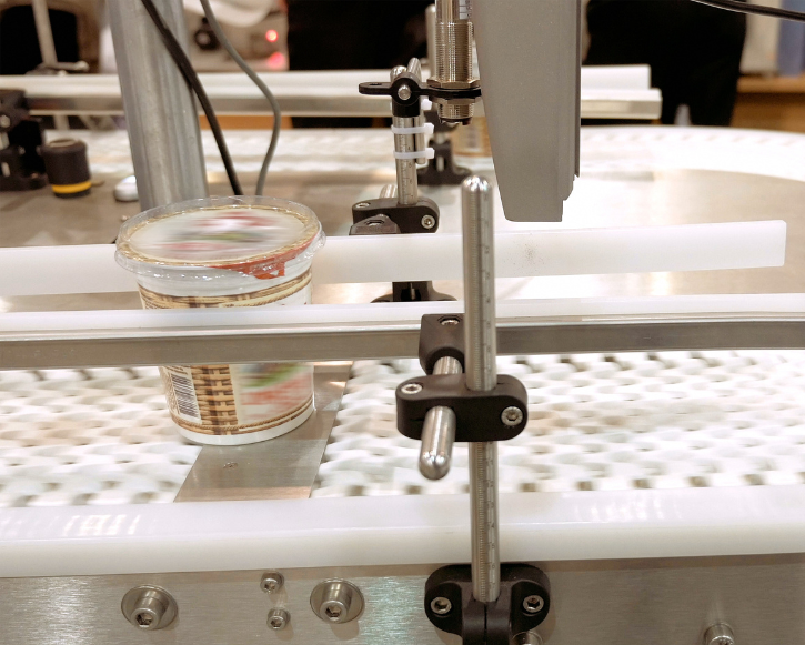 Plastic container with sealed lid on a metal conveyor belt, part of automated packaging machinery.