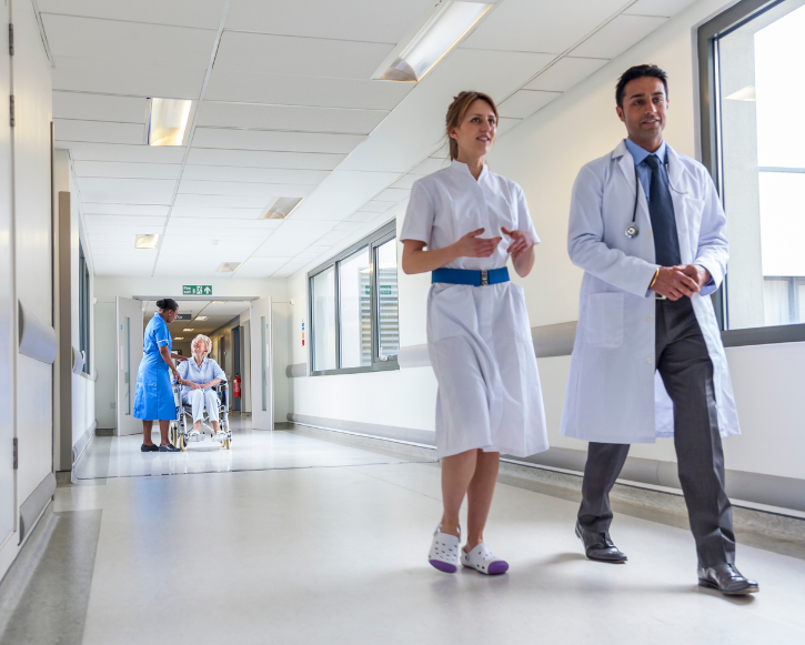 Two medical professionals walking down a brightly lit hospital corridor, with a nurse attending to an elderly patient in a wheelchair in the background.