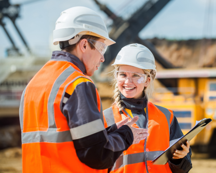 Two construction workers wearing white helmets and orange safety vests discussing and smiling, one holding a clipboard at a construction site.