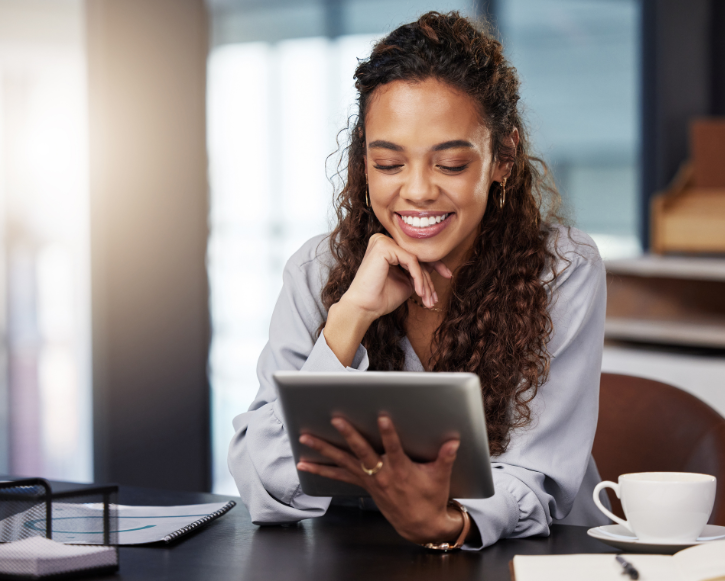 Smiling woman with curly hair looking at a tablet while sitting at a desk with a coffee cup and notebook.