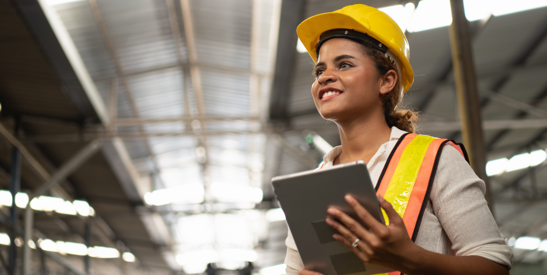 Smiling woman wearing a yellow hard hat and safety vest holding a tablet in an industrial warehouse.