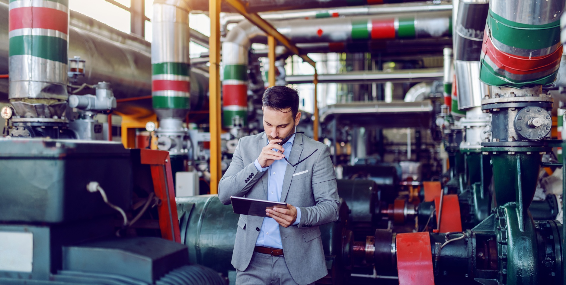 Man in a gray suit using a tablet inside an industrial plant with large machinery and pipes.