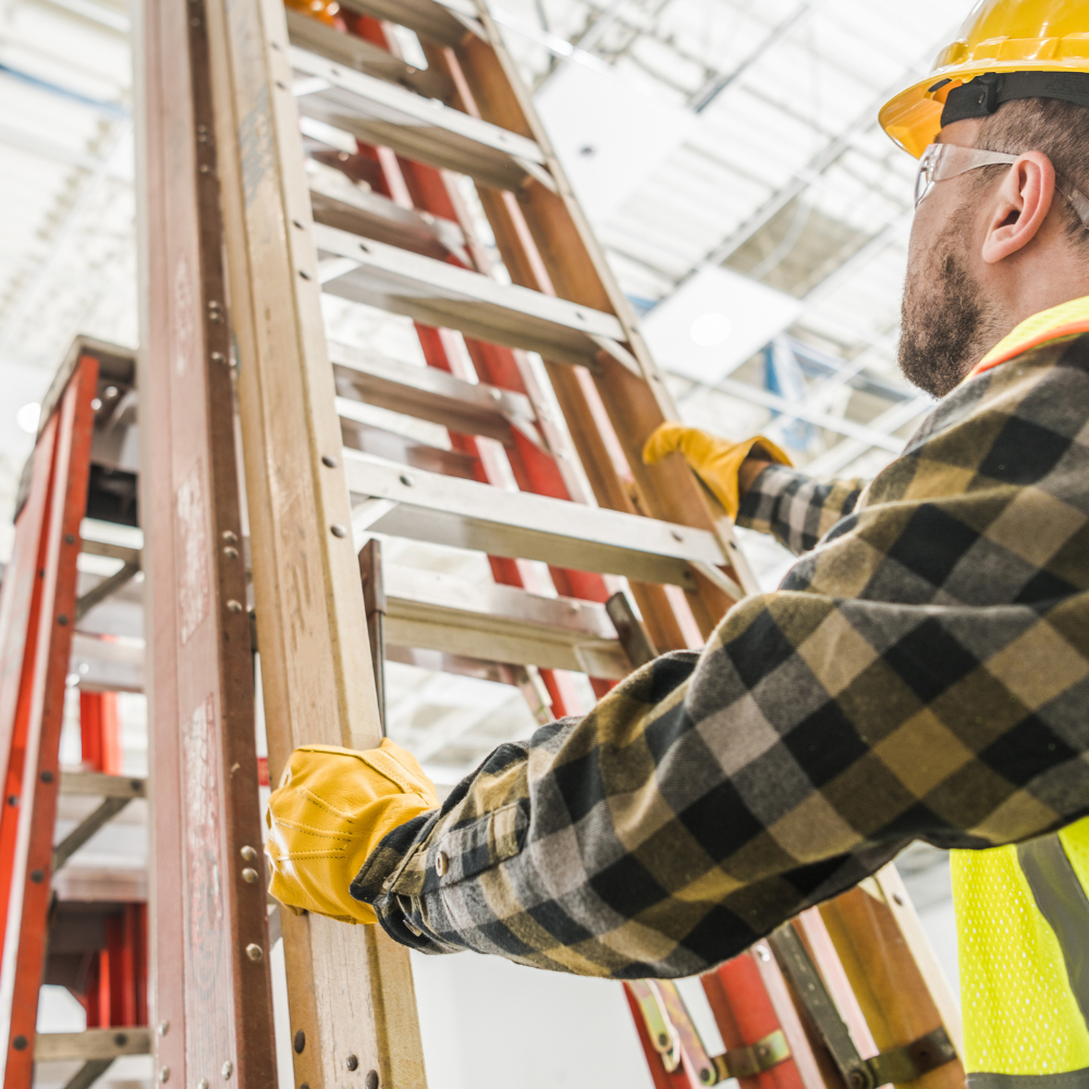 Construction worker in yellow hard hat and gloves holding a wooden ladder indoors.