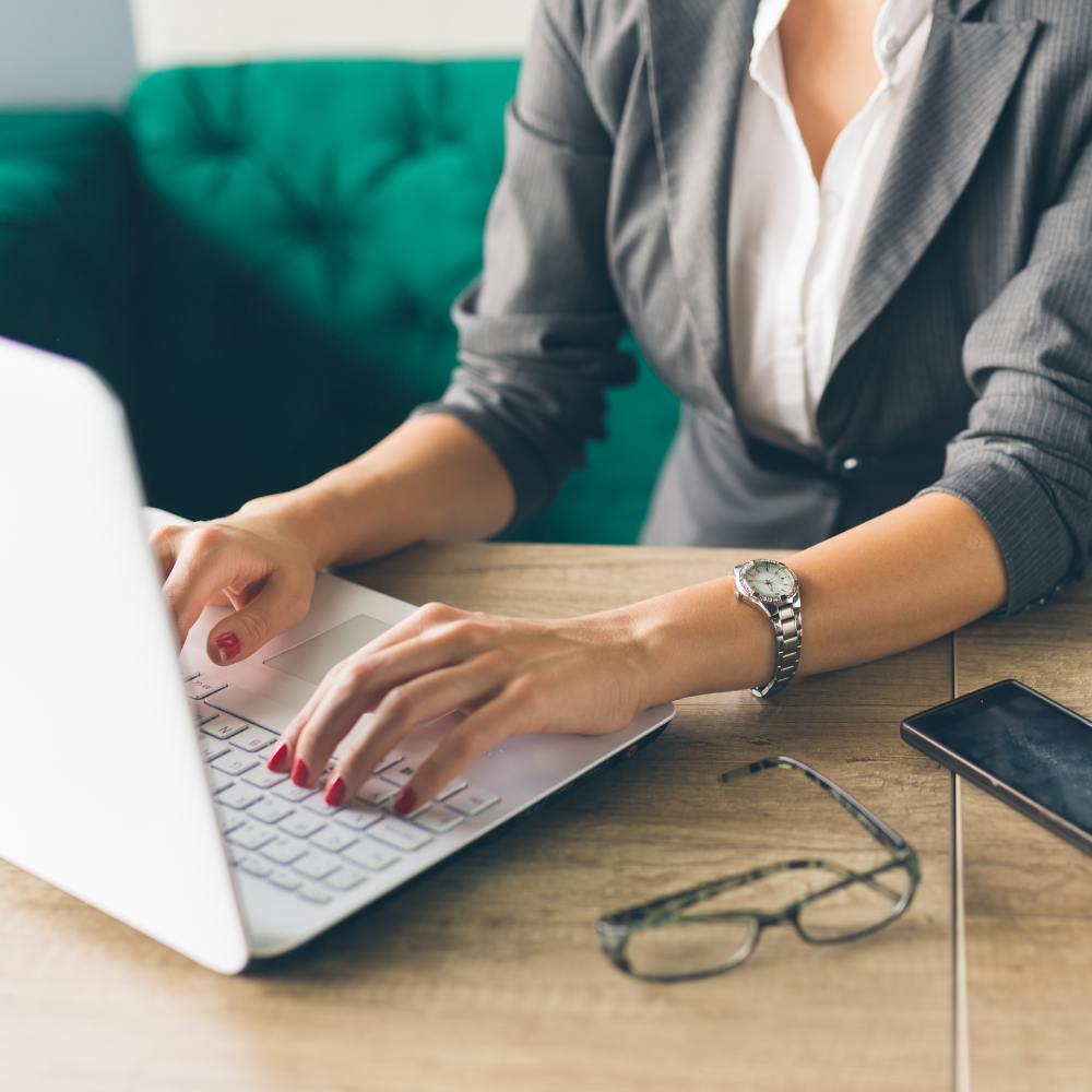 Woman in business attire typing on a laptop at a wooden desk with glasses and smartphone nearby.