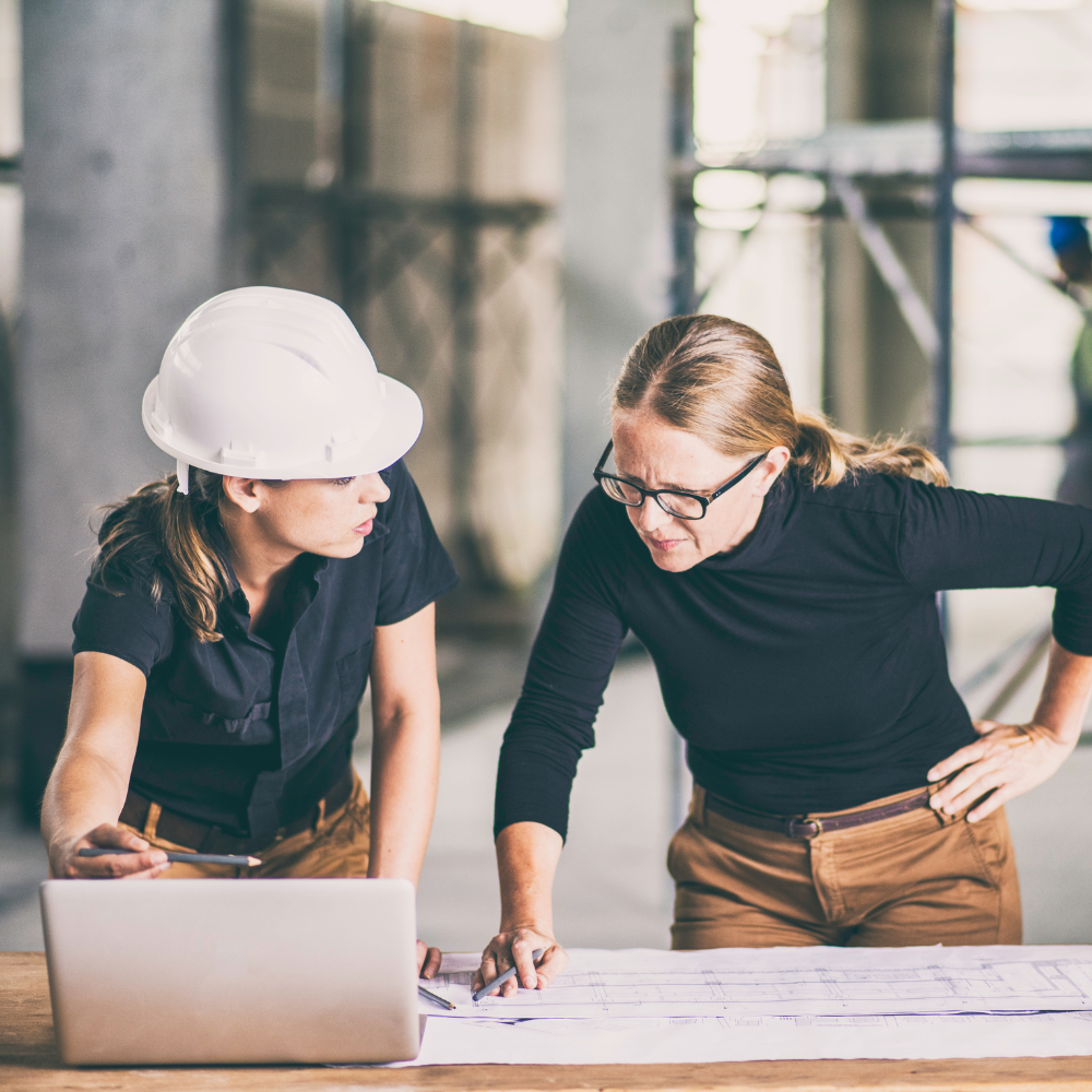 Two women reviewing blueprints on a table; one wearing a white hard hat and the other pointing at the plans.