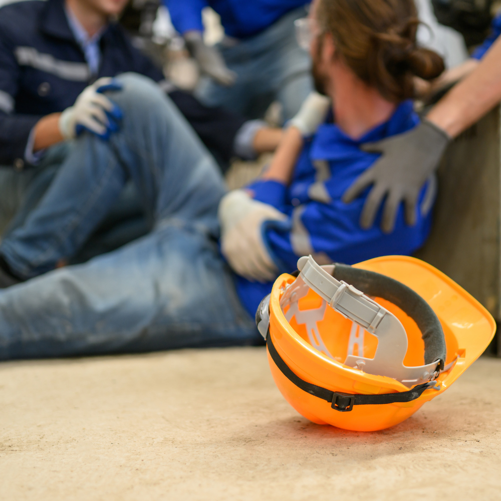 Orange safety helmet on the ground with blurred workers assisting an injured person in the background.
