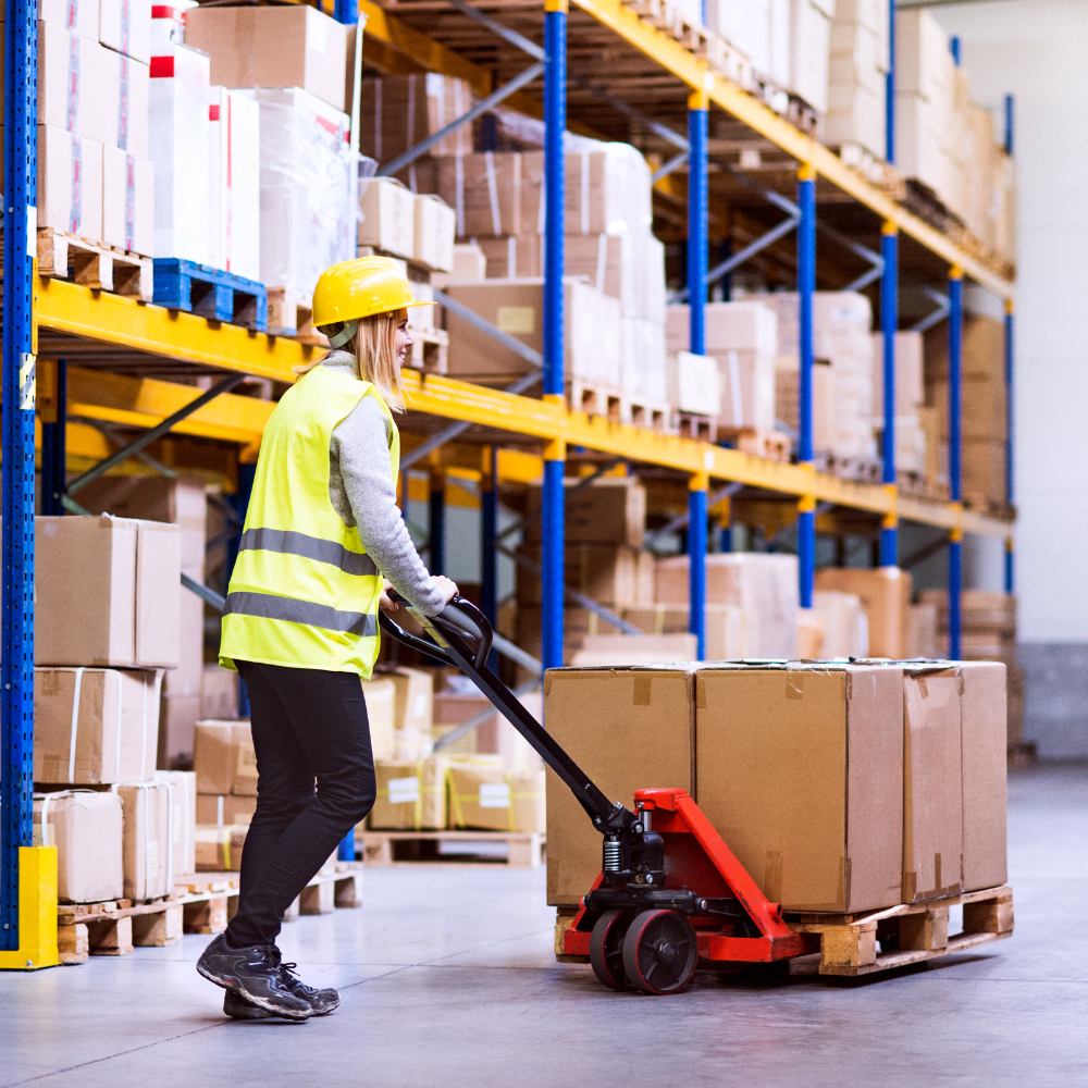 Warehouse worker wearing a yellow safety vest and hard hat using a pallet jack to move a loaded pallet of boxes.