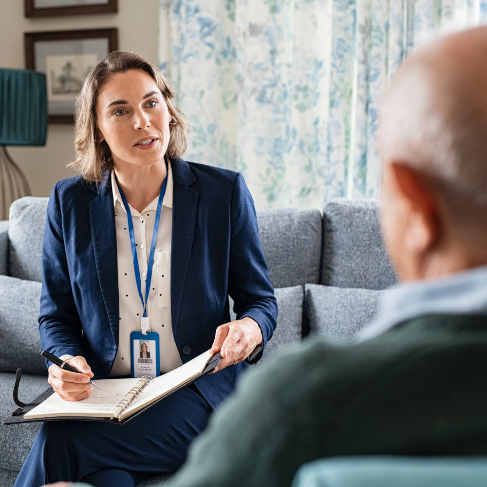 A female social worker in a navy suit attentively listens and takes notes while speaking with an elderly man in a home setting.