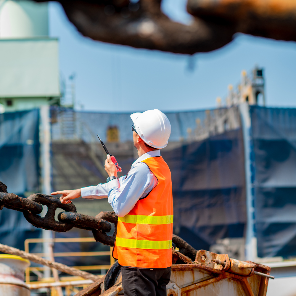 Construction worker in an orange safety vest and white helmet holding a walkie-talkie and pointing at a large chain.