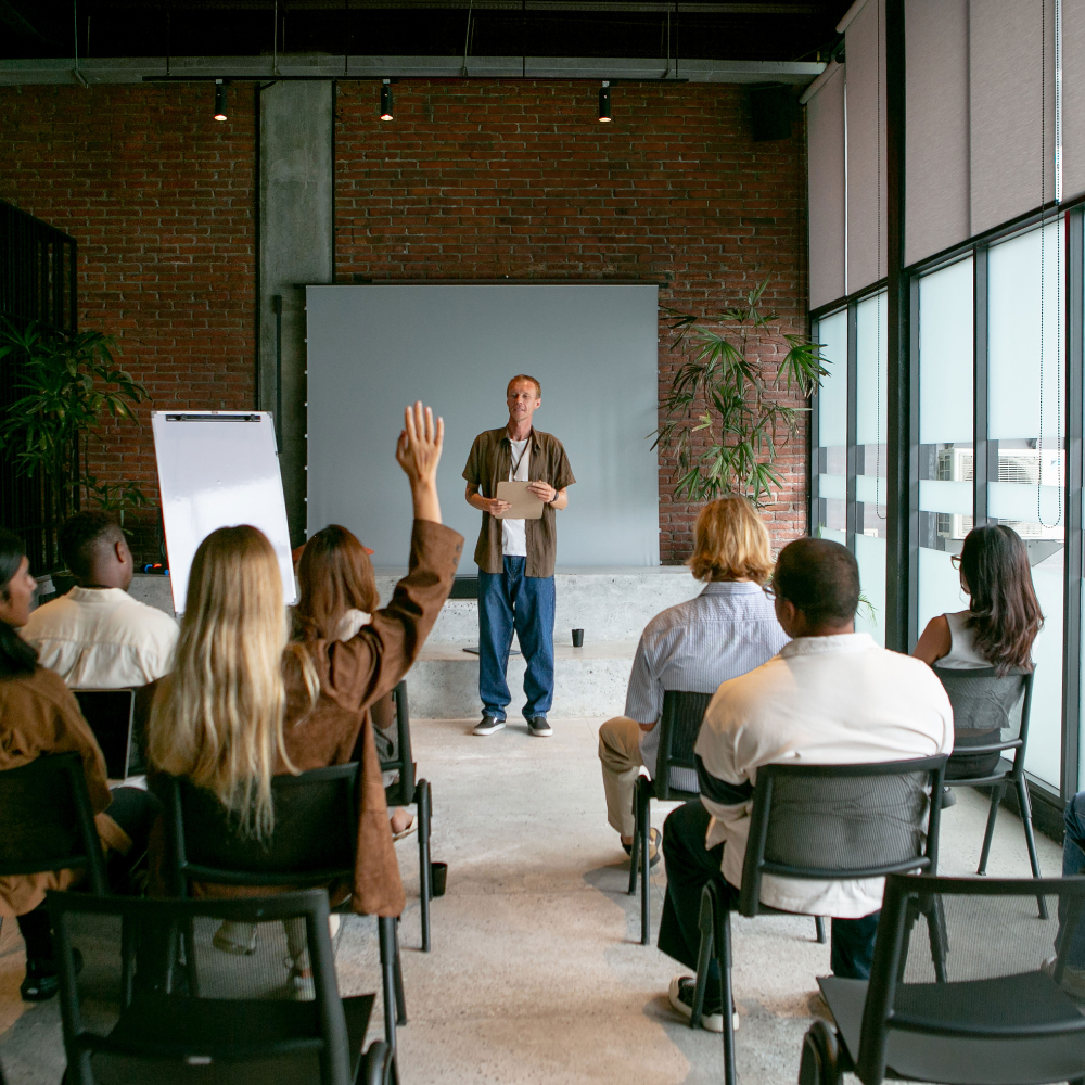 Man standing in front of a seated group in a modern room, one person raising their hand to ask a question.