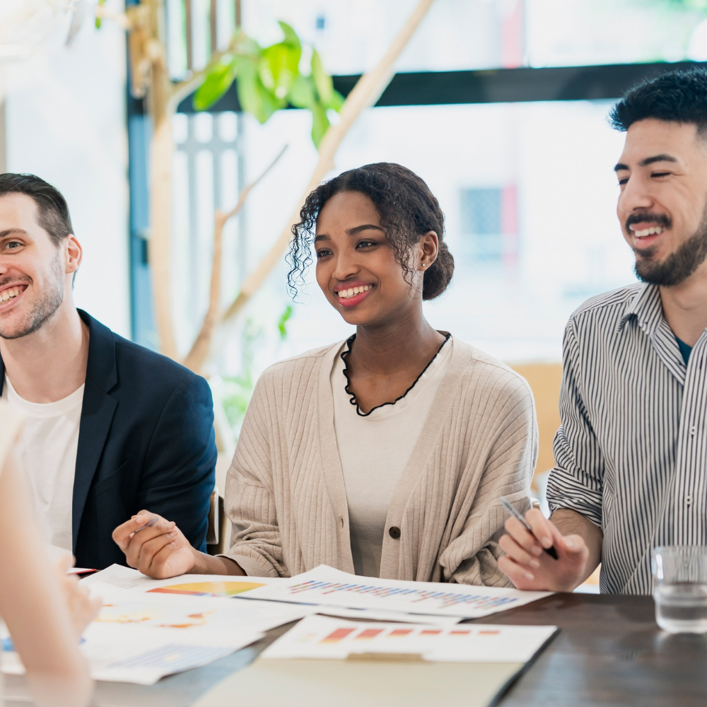 Three colleagues smiling and discussing charts and graphs at a meeting table in a bright office.