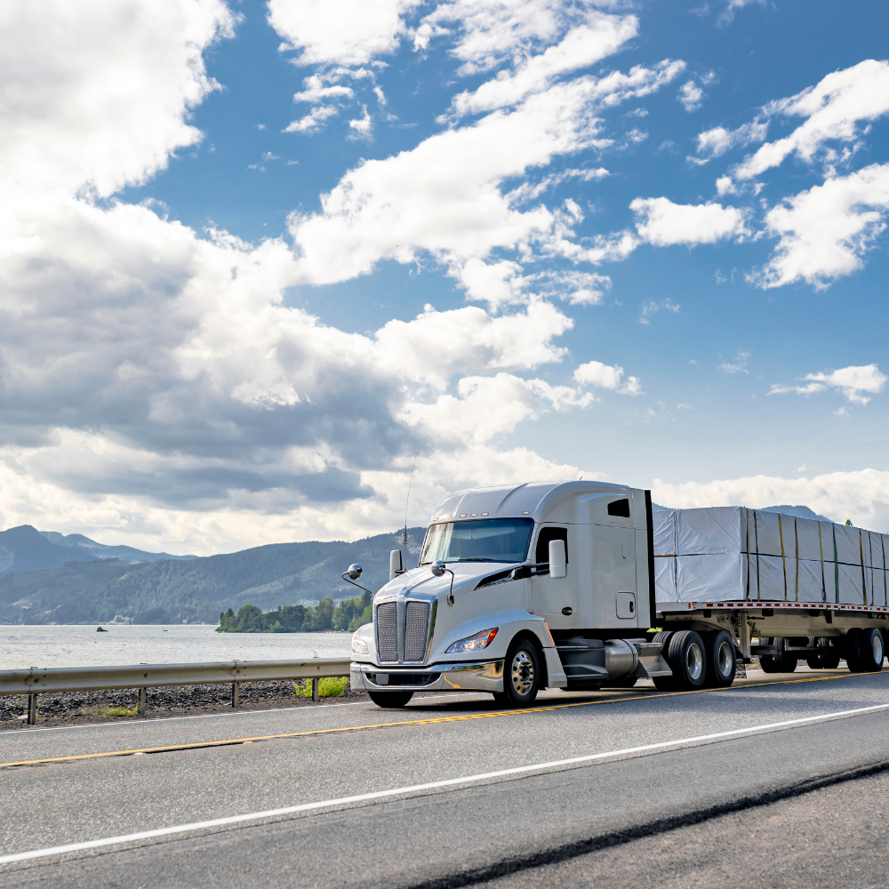 White semi-truck with covered trailer driving on highway beside a body of water and mountains under a partly cloudy blue sky.