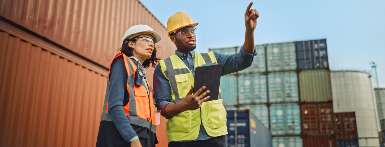 Two workers in hard hats and hi-vis vests at a container port, one pointing and one holding a tablet