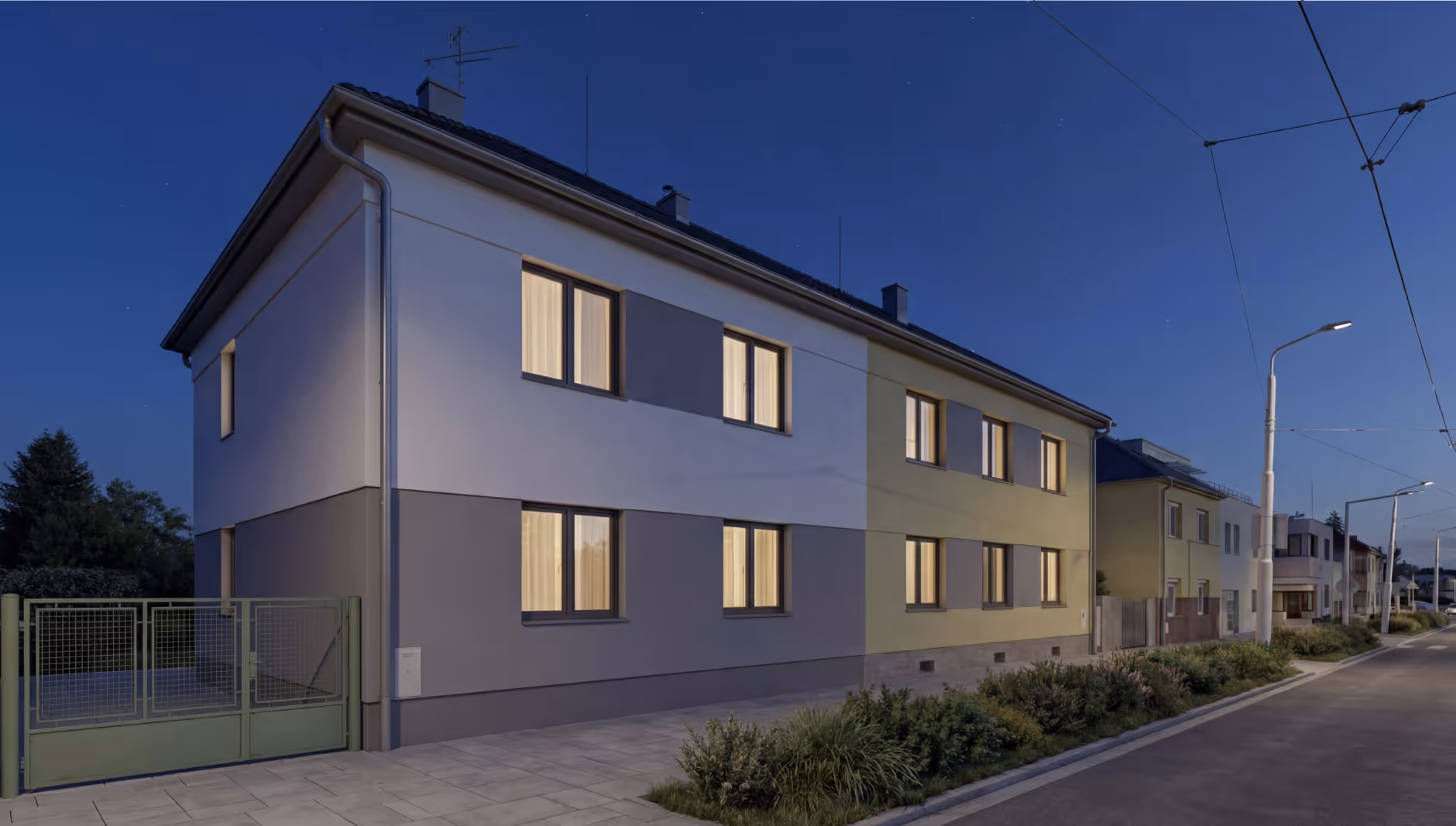 Dvoupodlažní rezidenční dům s osvětlenými okny večer na klidné ulici.