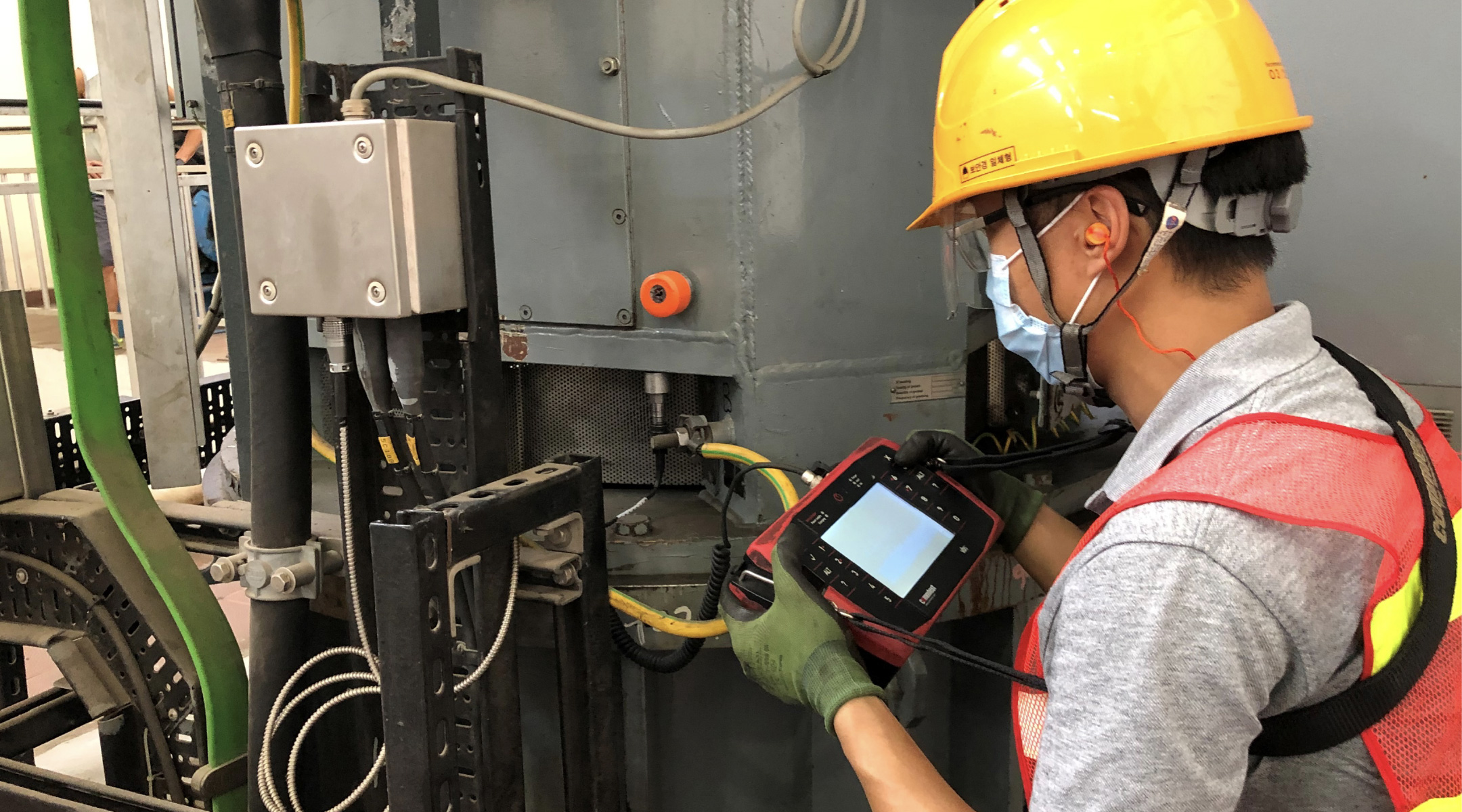 Technician wearing a yellow helmet and safety vest using a handheld diagnostic device to inspect industrial equipment.