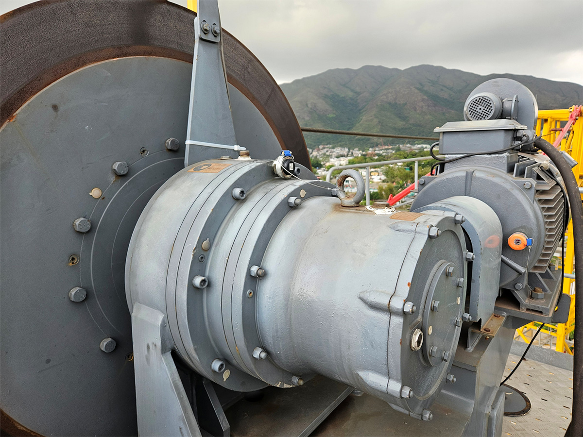 Close-up of industrial crane winch with large metal drum and motor, mountains visible in background.