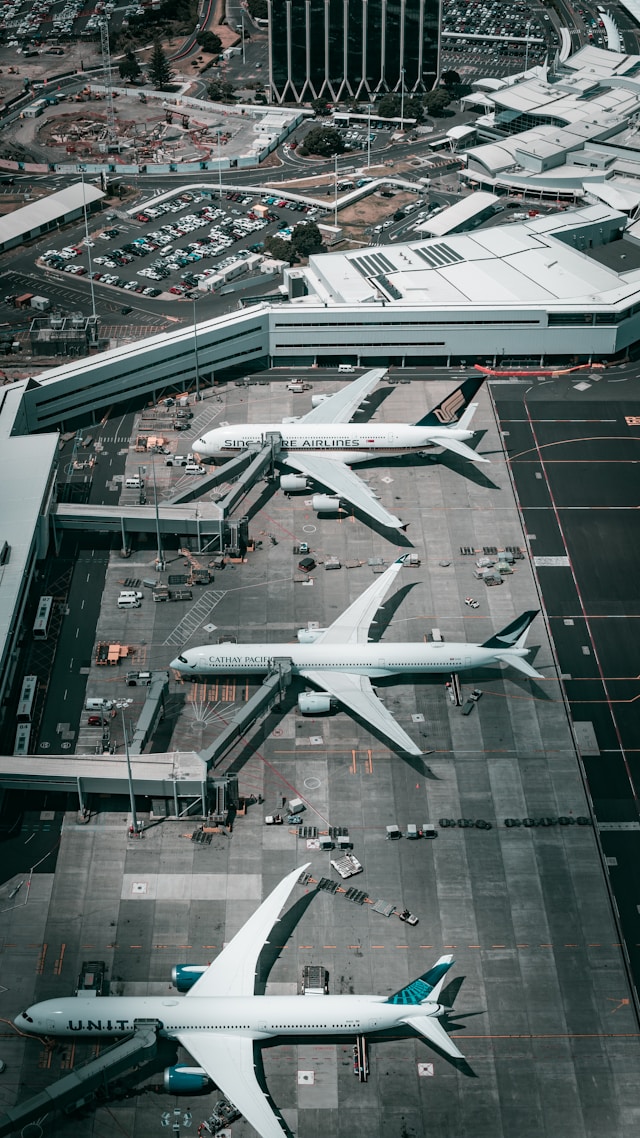 Aerial view of three airplanes from Singapore Airlines, Cathay Pacific, and United Airlines parked at airport gates with terminal buildings and parking lot in the background.