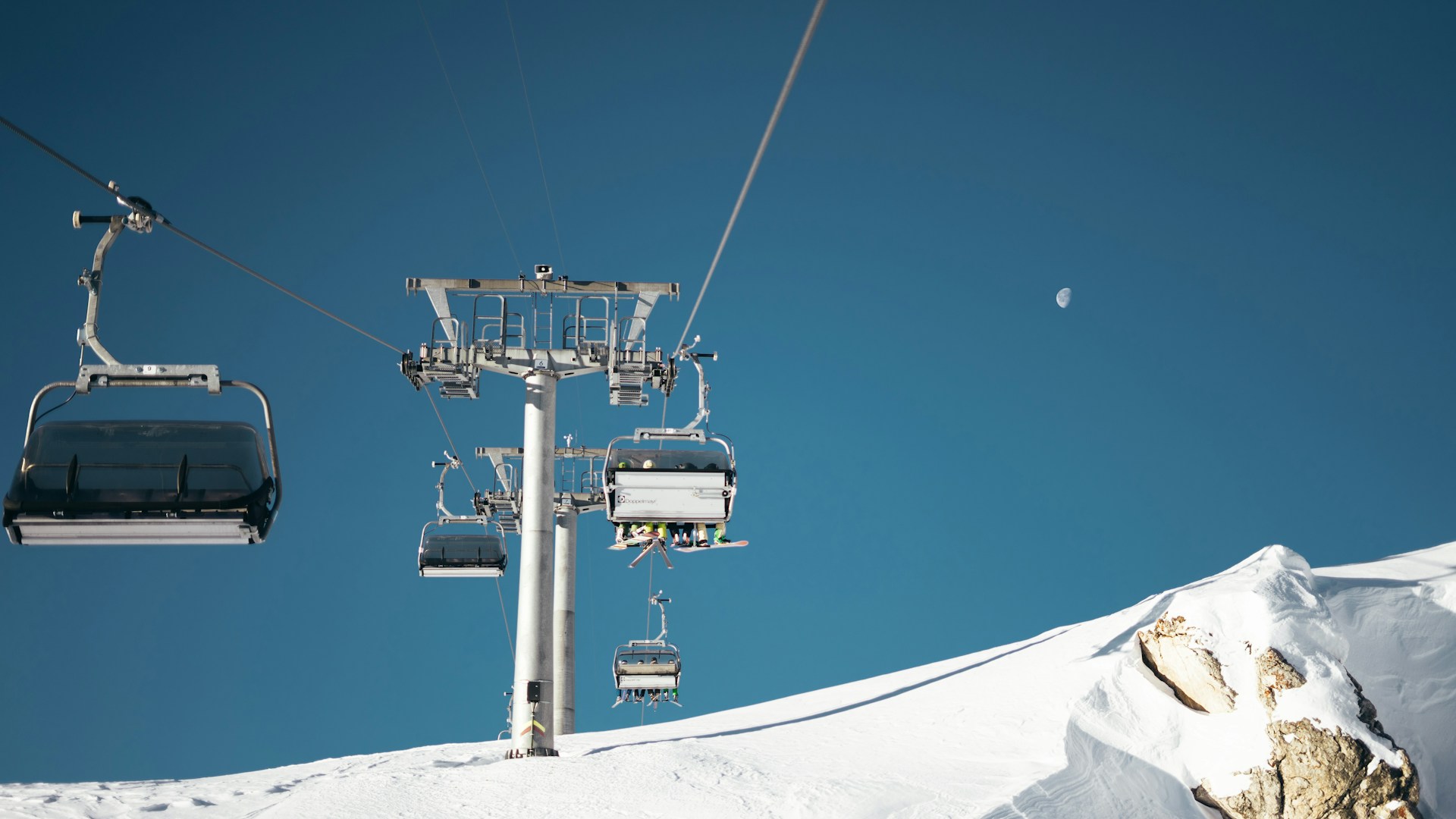Ski lift carrying people over a snowy mountain slope under a clear blue sky with a visible moon.