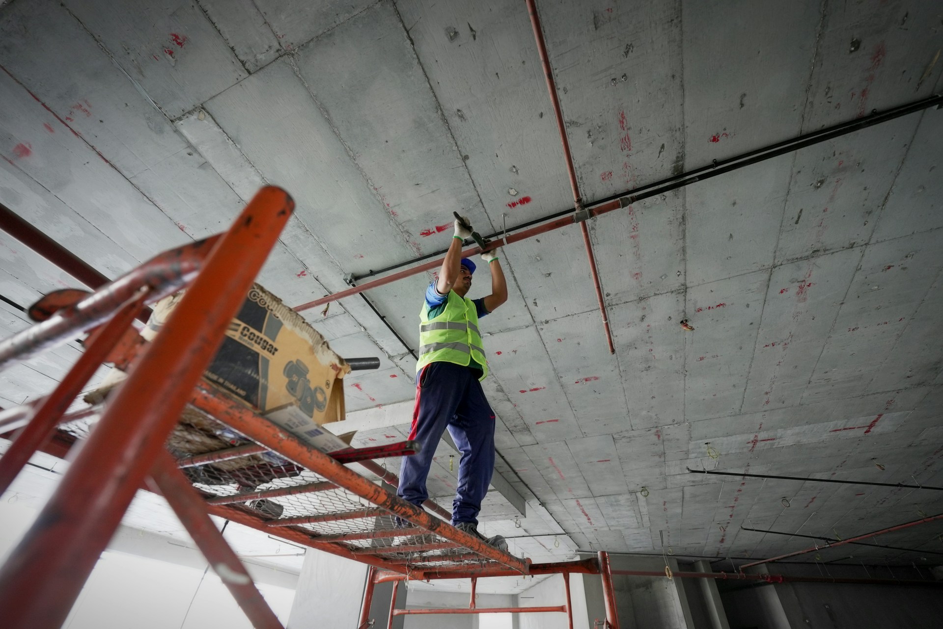 Construction worker wearing a safety vest and helmet standing on scaffolding while working on ceiling pipes.