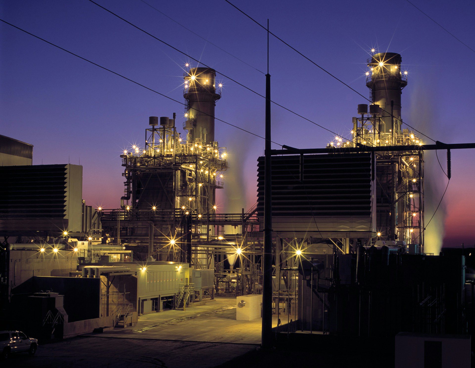 Industrial power plant at dusk with illuminated towers and steam emitting against a purple sky.