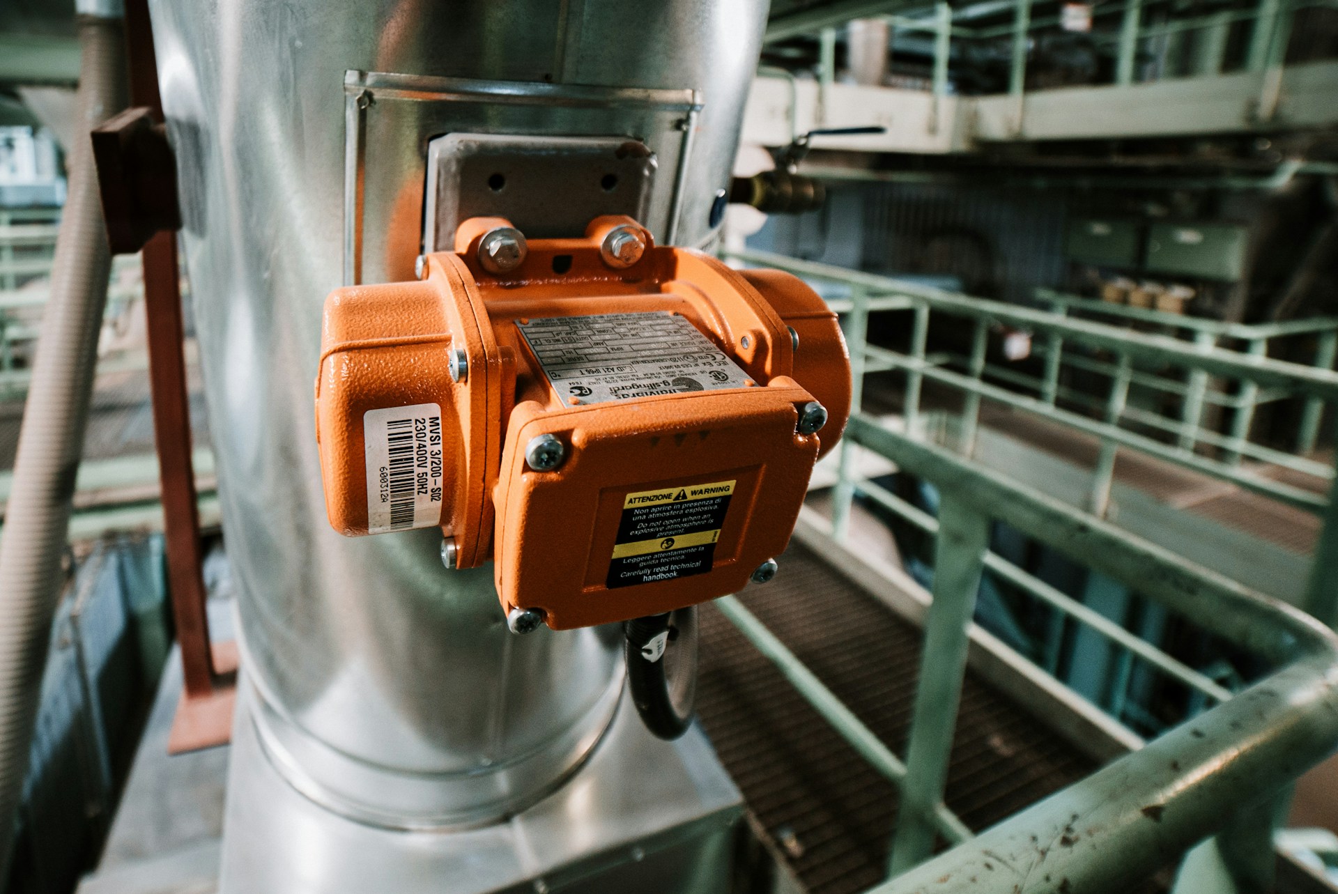 Close-up of an orange industrial electrical device mounted on a large metallic pipe inside a factory.