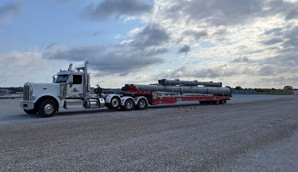White heavy-duty semi truck hauling large industrial pipes on a flatbed trailer under a cloudy sky.