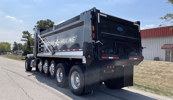 Black heavy-duty dump truck with 'Strong Hauling' logo parked on a road beside a building and grassy area.