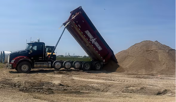 Dump truck unloading a large pile of sand or dirt at a construction site.