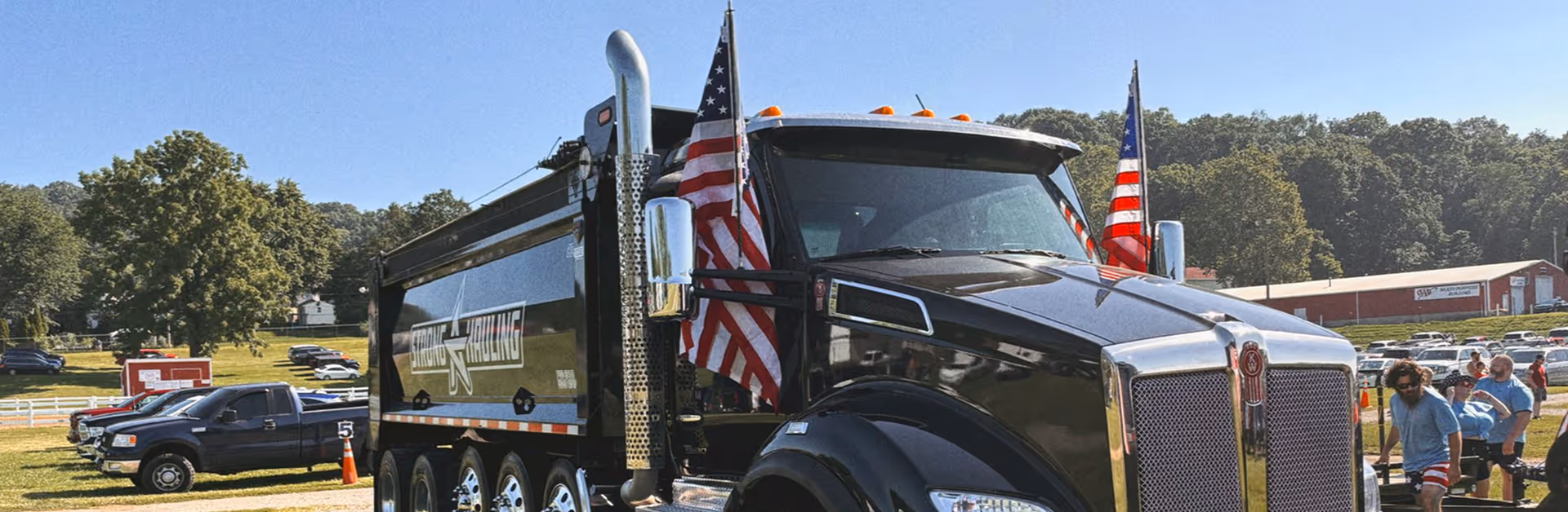 Black dump truck with American flags on front, parked outdoors near people and other vehicles on a sunny day.
