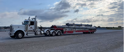 White semi-truck parked on gravel with a long flatbed trailer carrying large industrial pipes under a cloudy sky.