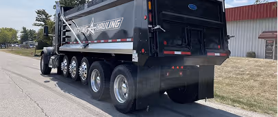 Black dump truck with multiple rear wheels parked on a road beside a grassy area and a building.