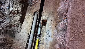 Two pipes exposed in a narrow trench with surrounding dirt and gravel.