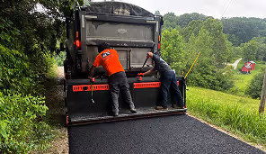 Two workers operating a large paving machine spreading black asphalt on a rural road surrounded by greenery.