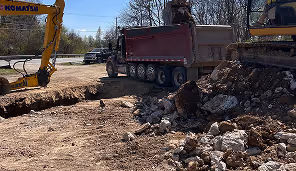 Construction site with excavators loading dirt and rubble into a large red dump truck near a trench.