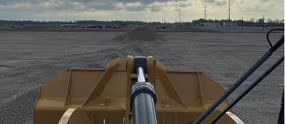 View over the bucket of a yellow excavator working on a large gravel or dirt surface near an industrial area.
