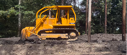 Yellow bulldozer with a front blade working on cleared soil near utility poles in a wooded area.